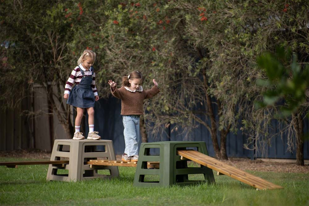 Two children balancing along a natural-timber trail between Step4 climbing blocks in a leafy garden